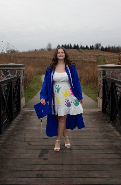 Early Childhood Education alum Alexis Nagel in dress with kids handprint paints on it and with graduation regalia on while standing on bridge that goes back to horizon focal point.