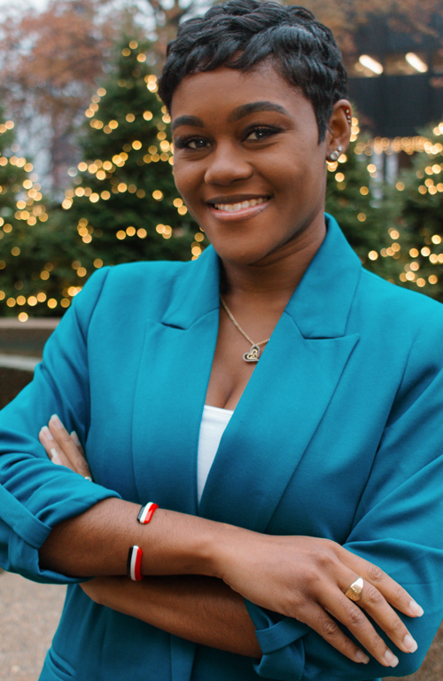 Fulbright Scholar and Ph.D. School Psychology student outside union smiling with arms crossed wearing blazer. and Christmas trees with lights in background.