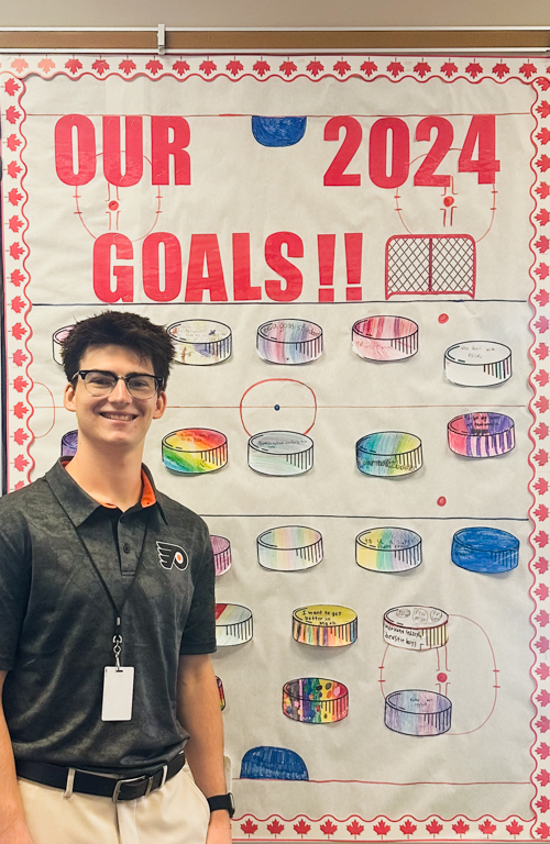 Evan Serakowski in classroom with bulletin board behind him that reads our 2024 goals!! and it has drawing of hockey rink on it with cut outs of colored hockey pucks that student wrote their goals on.