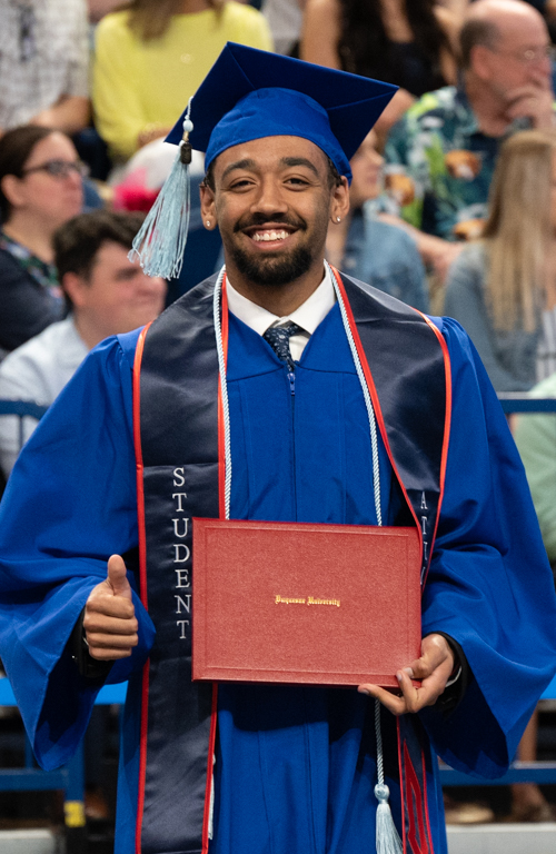 School of Education B.S.Ed. Secondary Education alum Ezekiel Daure smiles and gives a thumbs-up with diploma in-hand and commencement crowd behind him.