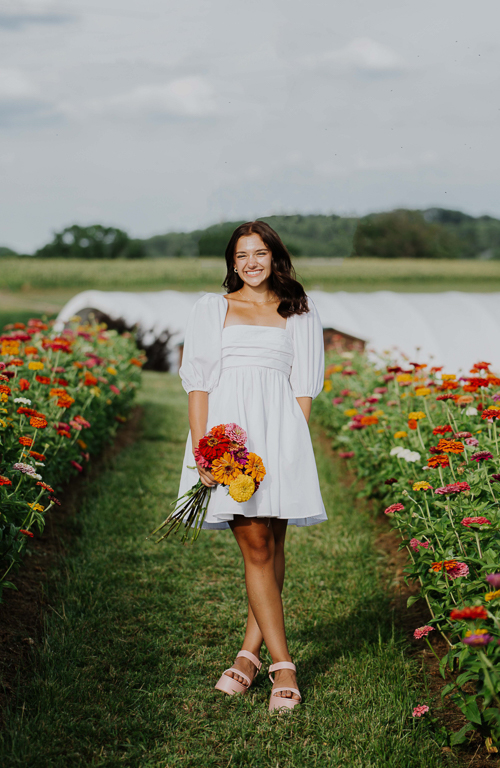 School of Education student Italia Cicione holding flowers with a field of flowers around her and a greenhouse and mountain behind her.