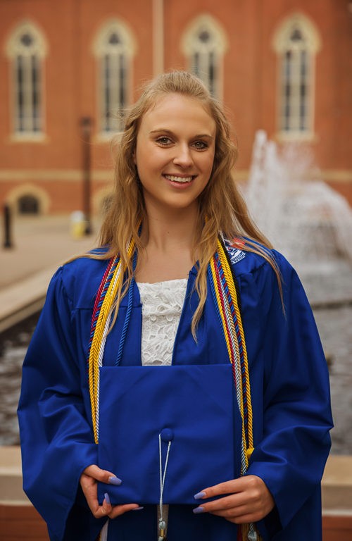Lauren Barnes in graduation regalia outside on campus with fountain and building behind her.