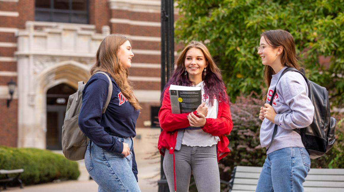 Three education students conversing on campus with Canevin Hall in background with bookbags and holding books.