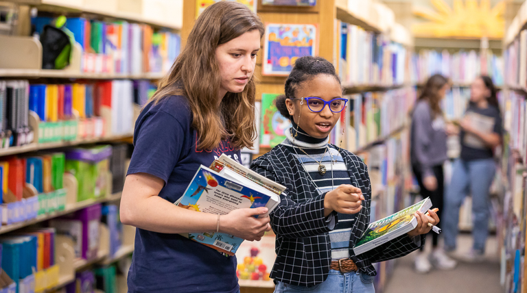 Two education students exploring resources in the library's curriculum center with books in hand and colorful bookshelves and student activity in the background.