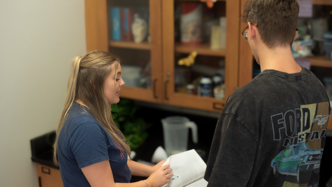 Student teacher in classroom reading science book standing with high school student.