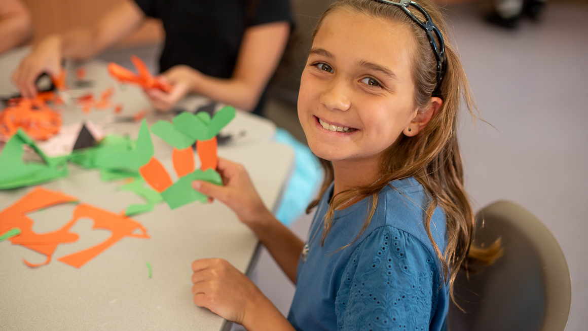 Ten-year-old Molly looking at viewer smiling with construction paper project in front of her at table in classroom.