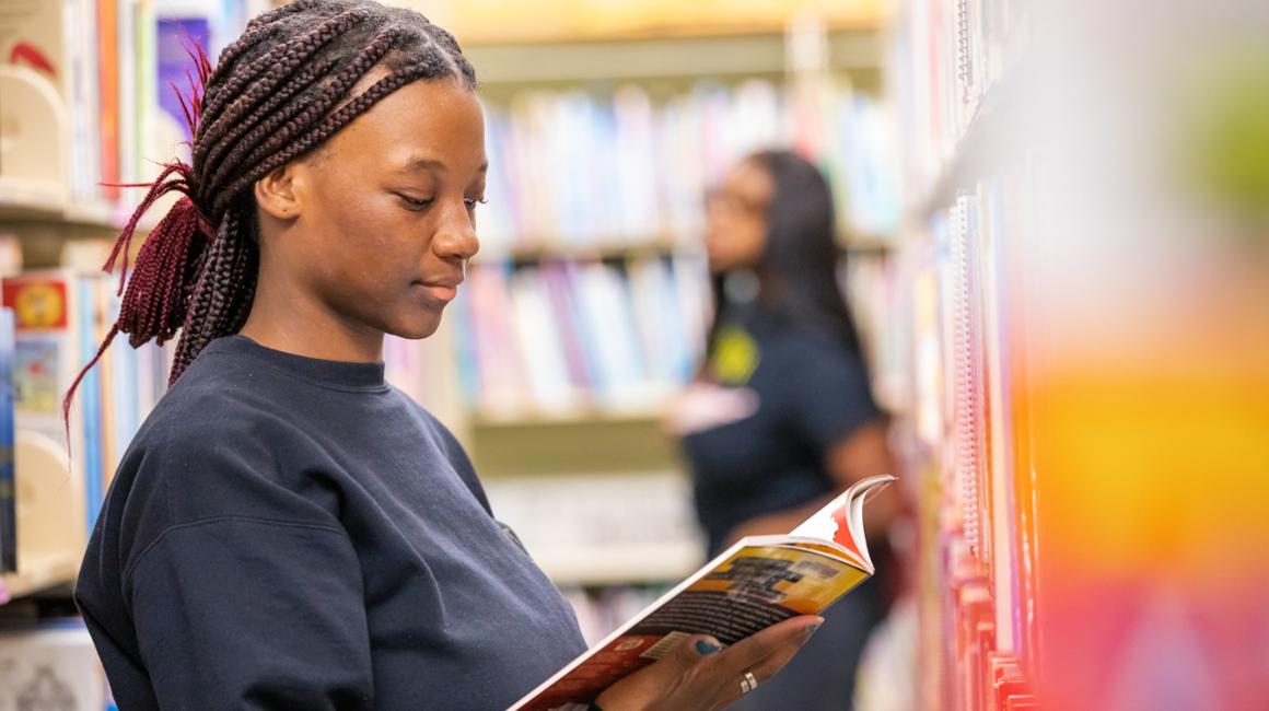 Education student in the library reading a book and another student walking in the background by bookshelves.