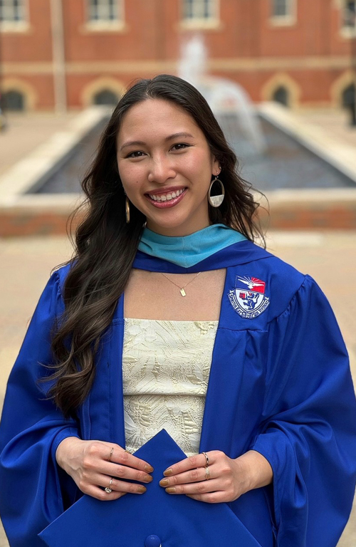 School of Education graduate Val Luutran smiles with graduation cap in hand dressed in graduation regalia in front of water fountain on Duquesne's campus.
