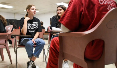 Two students seated in desks speak with an inmate.